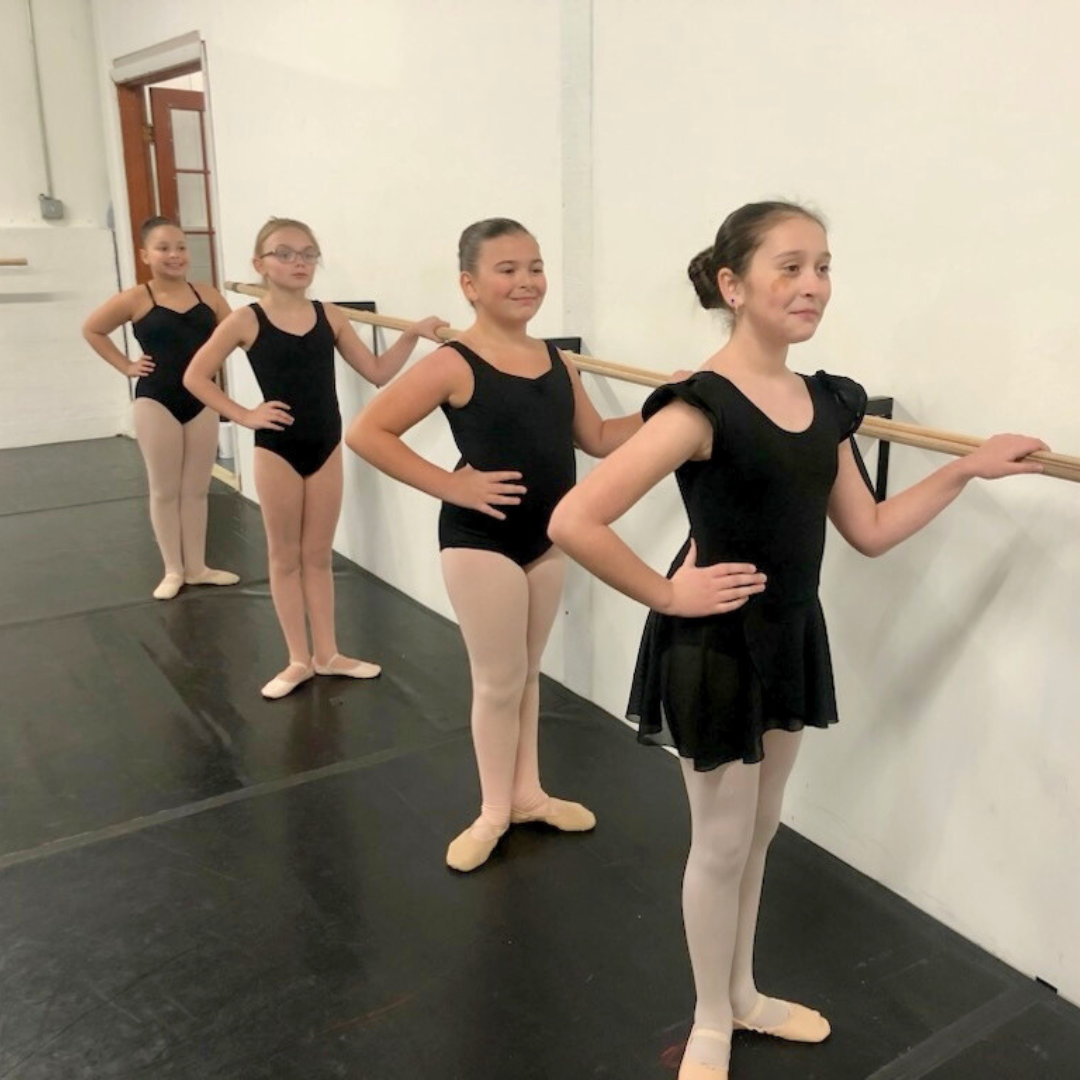 Four young dancers standing in first position at a ballet barre.