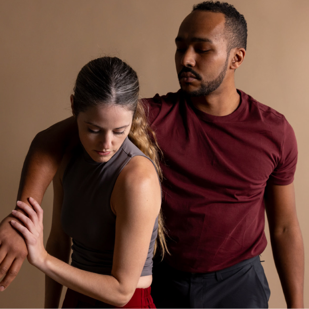 Two dancers from Boston Dance Theater, a woman on the right and a man on the left, stand and partially embrace in front of a beige backdrop.