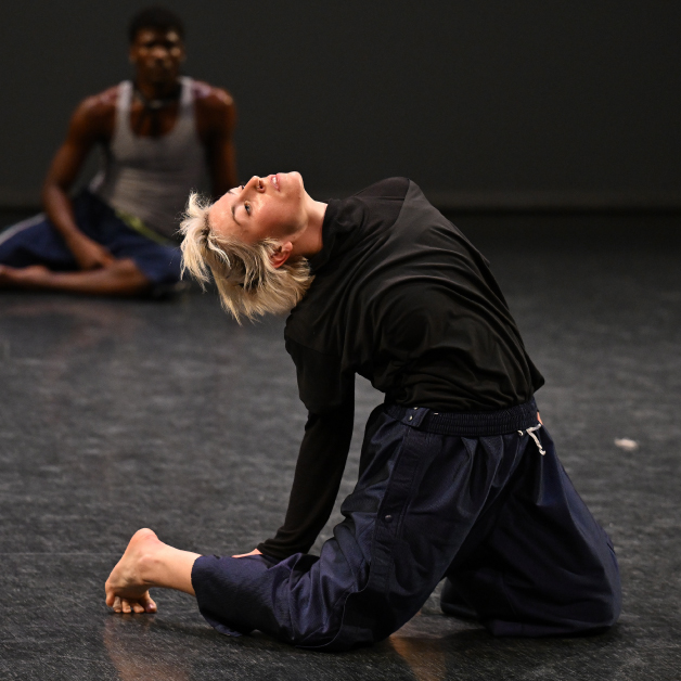 A dancer kneels atop a black floor, with another dancer sitting in the distance against a dark background.