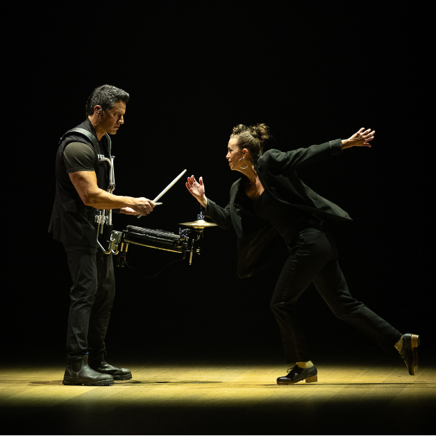 A dancer clad in black approaches a cymbal-player during a performance.