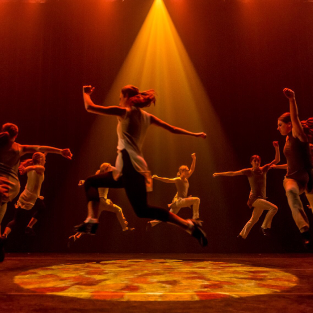 Members of Trinity Irish Dance Company jumping as a orange-hued projection lays on the floor beneath them.