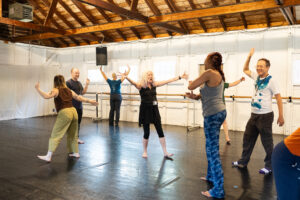 A group of people energetically engage in a dance workshop inside a studio with white walls and wooden beams.