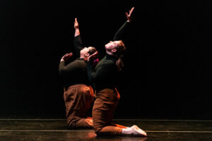 Two dancers dressed in black shirts and tan pants, pose on their knees, looking upwards toward one hand extended.