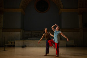 Two dancers perform in a dimly lit hall with arched ceilings. One holds a poised stance with a raised arm, while the other follows, creating a dramatic scene.