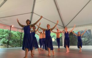 A group of dancers in blue raise their arms while standing under a white tent