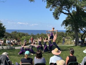 A crowd of people in lawn chairs watch as dancers in body-tight suits tilt their bodies towards the sky; one dancer is held up by two others