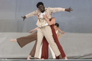 A dancer in a tan suit set stands with arms stretched to the sides. Another dancer grabs them from behind.
