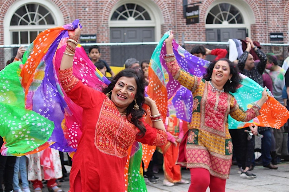 Two dancers move joyfully in bright embroidered outfits, lifting colorful scarves overhead in rhythmic, sweeping gestures. Their bodies twist and extend with expressive energy, surrounded by a lively crowd sharing in the festive dance.